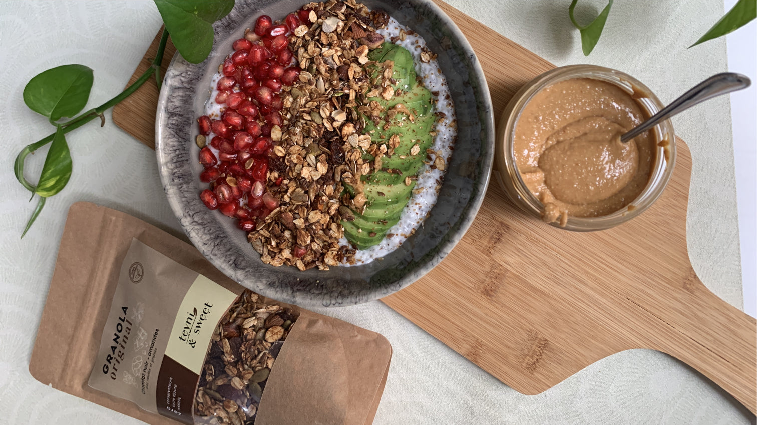 Bol de petit-déjeuner healthy avec yaourt, granola croustillant, avocat tranché et graines de grenade, accompagné d’un pot de beurre de cacahuète et d’un sachet de granola Teyni & Sweet.