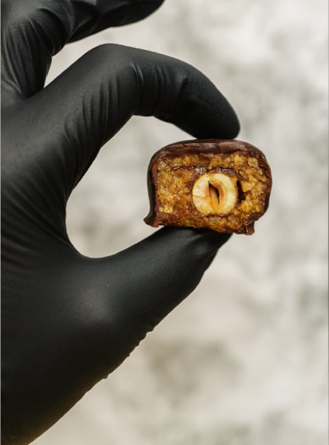 Boule d’énergie coupée en deux, montrant un cœur de noisette entière, tenue entre deux doigts gantés.