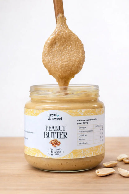 Beurre de cacahuète - peanut butter artisanal Teyni & Sweet dans un pot en verre, avec une cuillère en bois plongeant dans une texture onctueuse, posé sur une planche en bois avec des cacahuètes autour.