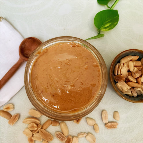 Vue du dessus d’un pot de beurre de cacahuète artisanal Teyni & Sweet, texture lisse et naturelle, entouré de cacahuètes entières et d’une cuillère en bois sur fond clair.