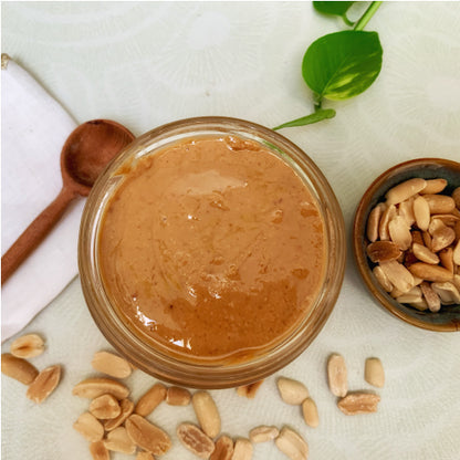 Vue du dessus d’un pot de beurre de cacahuète artisanal Teyni & Sweet, texture lisse et naturelle, entouré de cacahuètes entières et d’une cuillère en bois sur fond clair.